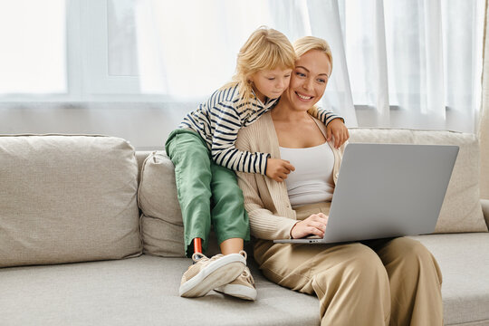 happy girl with prosthetic leg hugging blonde mother working on laptop in modern living room