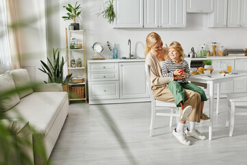 little girl with prosthetic leg sitting on laps of happy blonde mother during breakfast in kitchen