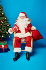 happy Santa Claus in eyeglasses sitting in rocking chair with gift and sack bag near Christmas tree