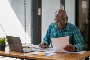 A senior black man in casual clothes is busy at work but happy with the work and paperwork in front of him.