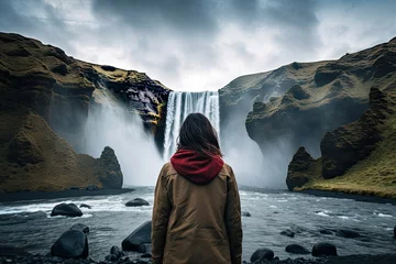 Deurstickers A young woman in a yellow jacket and a red scarf is standing in front of the Skogafoss waterfall in Iceland, rear view of a Woman overlooking a waterfall at skogafoss, Iceland, AI Generatedv © Iftikhar alam