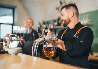 Fresh lager beer glass mug and barman dressed uniform smiling to teammate waiter with tray beer...