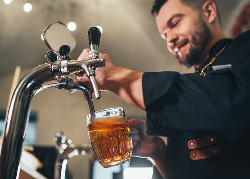 Tapping fresh lager beer in glass mug close up. Smiling stylish bearded barman dressed black uniform with an apron at bar counter. Successful people, beer consumption, beverages industry concept imag