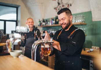 Stylish bearded barman dressed black uniform smiling at camera, beer tapping at bar counter and waiter with tray. Successful people teamwork, friendship, brewing and restaurant industry concept image