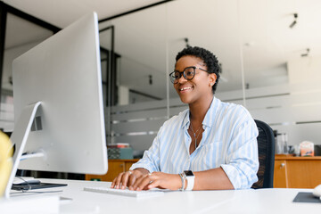 Obraz na płótnie Canvas Successful african-american woman sitting at desk with laptop, smiling and typing, enjoying office job, female employee , businesswoman feeling herself on the right place