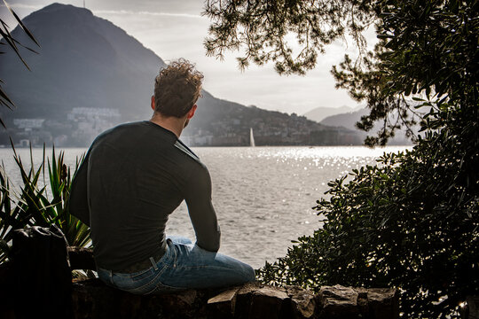 A man sitting on a rock looking out over a body of water in Lugano, Switzerland - Powered by Adobe