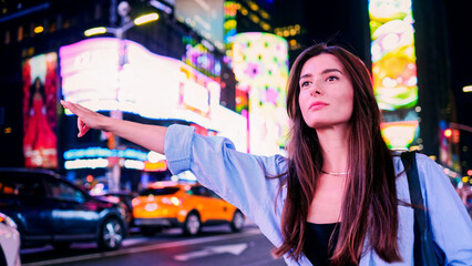 Young beautiful charming female woman waving taxi in New York City at night. Attractive tourist walking in background of pedestrian crossing, city center. Taxi car traffic bokeh effect.