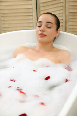Woman taking bath in tub with foam and rose petals indoors
