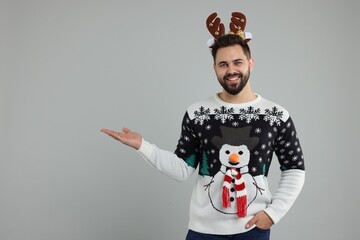 Young man in Christmas sweater and reindeer headband showing something on grey background. Space for text