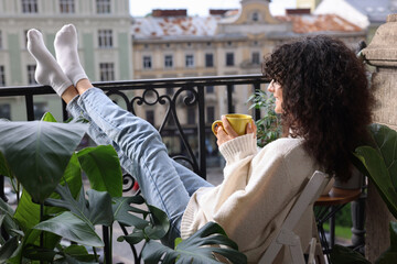 Young woman with cup of tea relaxing in chair surrounded by green houseplants on balcony - obrazy, fototapety, plakaty