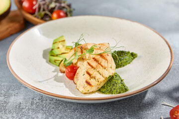 Horizontal side view of a grilled chicken fillet dish with avocado and spinach on a ceramic plate. Set against a grey concrete background with hard shadows for a modern culinary appeal
