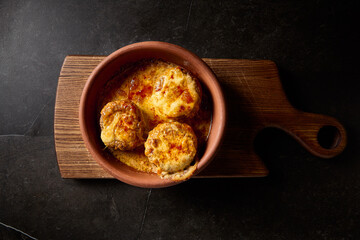 Horizontal top view of baked mushrooms filled with suluguni cheese, presented on a traditional Georgian 'keci' clay plate, placed on a wooden board with a contrasting black background