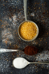 Spoons with sugar and ground coffee on black background sprinkled with sugar, coffee, cocoa powder.