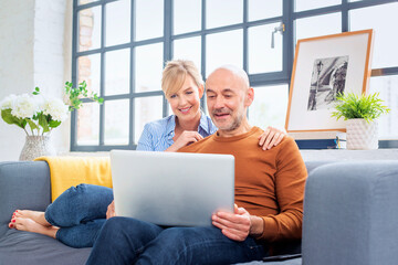 Shot of a blond haired woman and mid aged man sitting on the sofa at home and using laptop