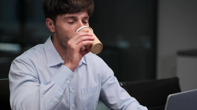 Man pauses to take a sip of hot beverage, finding brief respite amidst the tasks on laptop. The steam rises from cup, momentarily veiling focused expression.