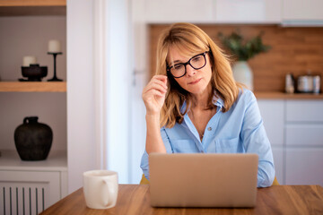 Thinking mid aged woman sitting at home and using laptop for work or having video call