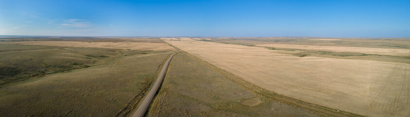 Aerial panoramic view of an extensive dry prairie landscape under a bright blue sky. A rough looking road crosses the fields and disappears into the horizon.
