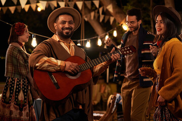 Happy mexican man playing guitar during traditional holiday with people dancing in background