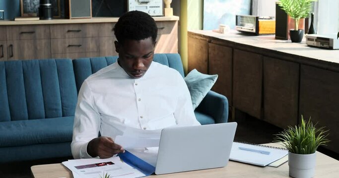 An organized African American man manages tasks from home, juggling papers and laptop with precision. His dedication and multitasking skills shine as he handles work responsibilities.