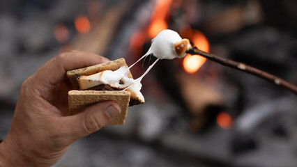 Closeup hand roasting marshmallow enjoying s'more by a campfire. Camping cooking concept.	
