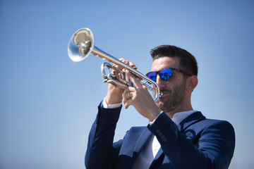 Young Hispanic man, wearing a jacket and sunglasses, playing a pretty, silvery trumpet outdoors....