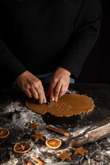 Top view of woman's hands with mold on gingerbread cookie dough on dark wooden table with cinnamon, flour, oranges and rolling pin, vertical