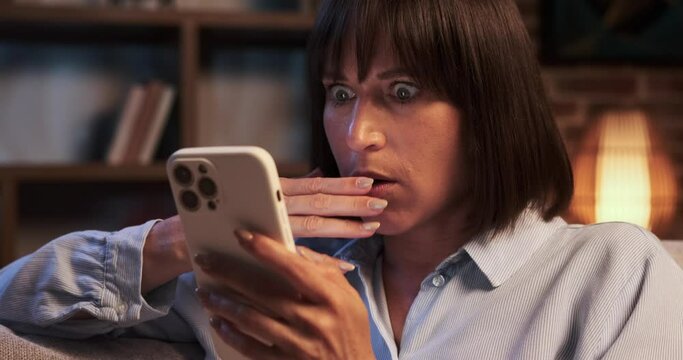 Startled Caucasian woman clutches phone tightly in the living room, eyes wide with fear. The room seems to echo her sense of unease, the atmosphere tense and apprehensive.