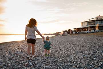 Young mother spends time with cute baby child toddler by the sea at sunset