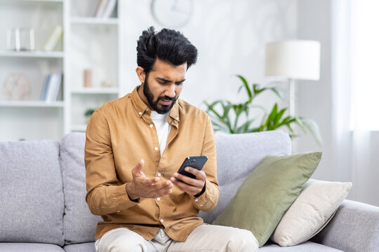 Sad unhappy and disappointed man sitting at home on sofa in living room, Indian man received notification message with bad news, reading using smartphone app.