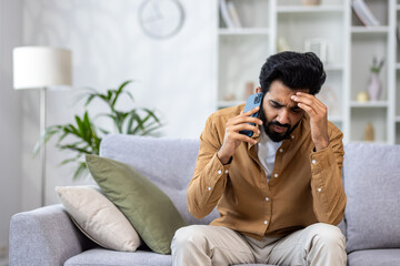 Angry angry man talking on the phone at home, dissatisfied customer sitting on the sofa in the living room yelling at the interlocutor.