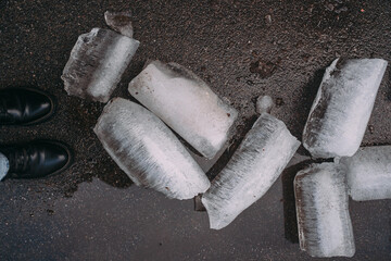Women's feet in black boots on the sidewalk. There are blocks of ice on the sidewalk that have fallen from the drainpipe and roof. Icicles are lying on the asphalt - obrazy, fototapety, plakaty