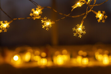 Stylish glowing snowflake christmas garland on background of golden lights bokeh in evening cozy room. Festive illumination. Christmas banner, copy space. Merry Christmas and Happy Holidays!