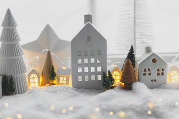 Naklejka premium Cozy christmas miniature village. Stylish cute little ceramic houses and wooden trees on soft snow blanket with glowing lights in room. Atmospheric winter village still life. Merry Christmas!