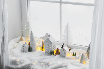 Atmospheric miniature winter village. Stylish cute little ceramic houses and christmas wooden trees on soft snow blanket with glowing lights in room. Christmas modern white background.