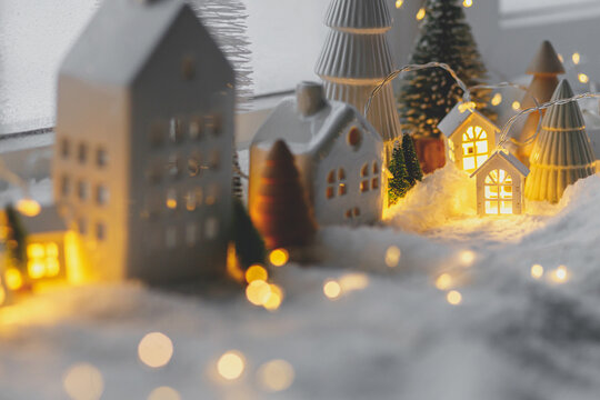 Cozy christmas miniature village. Stylish little ceramic houses and wooden trees on soft snow blanket with glowing lights in evening. Atmospheric winter village still life. Merry Christmas!