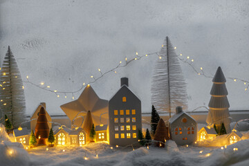 Cozy christmas miniature village. Stylish little ceramic houses and wooden trees on soft snow blanket with glowing lights in evening. Atmospheric winter village still life. Merry Christmas! - obrazy, fototapety, plakaty