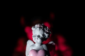 A toy white angel with heart in his hands against the background of a huge red demon with horns