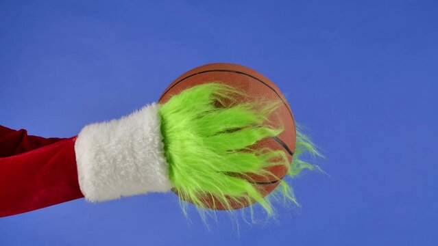 Grinch's green hairy hands holding out a basketball on blue isolated background. Gift snatcher cosplay. Christmas and new year celebration concept. Blue screen, chroma key.