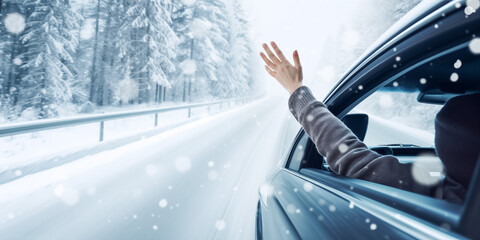 Happy woman in car waving while driving through winter forest. Woman driver feels wind through her hands while driving in winter landscape. Winter vacation and freedom concept

