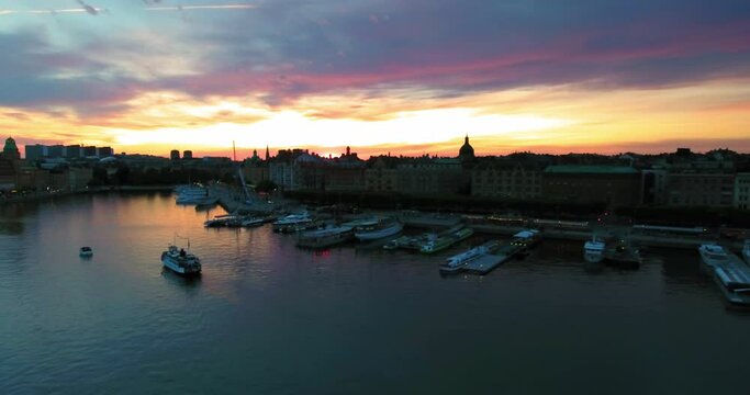 Aerial Backward Ascending Shot Of Boats In Sea By Buildings At Sunset Against Cloudy Sky - Stockholm, Sweden