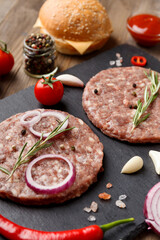 Raw beef hamburger patties on stone plate, vegetables and spices, wooden background.