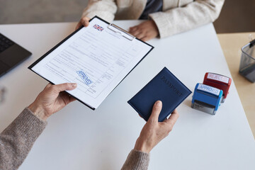 Above angle of female manager passing approved document and passport to applicant over desk after stamping visa application form