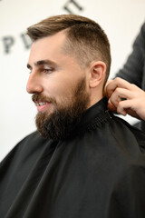 A young man with a beard in a black cape sits on a chair in a barbershop. The client is waiting for a haircut of the beard.