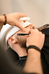 A barber shaves the cheek of a bearded customer with dangerous razor. Shaving the contour of the beard for the correct shape.