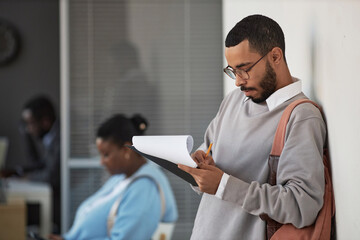 Young male applicant in grey pullover and eyeglasses filling in visa application form and writing...