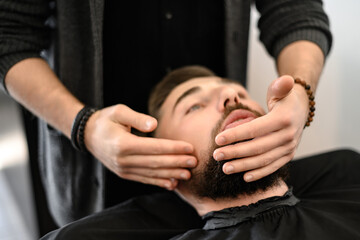 A barber massages and rubs a male client beard after applying moisturizing oil - obrazy, fototapety, plakaty