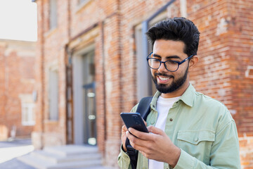 Portrait of young successful student outside university campus, man smiling and using app on phone, typing message and browsing social media, indian man holding phone with backpack - obrazy, fototapety, plakaty