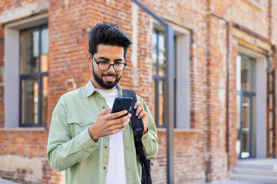 Portrait of young successful student outside university campus, man smiling and using app on phone, typing message and browsing social media, indian man holding phone with backpack