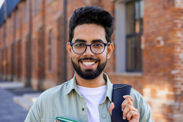 Portrait of young successful smiling student, Indian man close up smiling and looking at camera, man standing outside university campus in casual clothes with backpack and books in hands