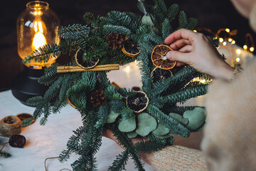 Young woman in knitted sweater doing handmade Christmas wreath from natural materials: fir tree branches, dry citrus slices, cones, cinnamon sticks. Wooden table, cozy light, home atmosphere